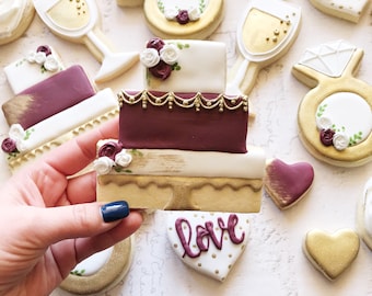 Puede incluir: Surtido de galletas de azúcar decoradas en varias formas, incluyendo una tarta de tres pisos, copas de champán, un anillo de diamantes y corazones. Las galletas presentan glaseado dorado, blanco y burdeos con detalles florales. Una galleta dice "love".