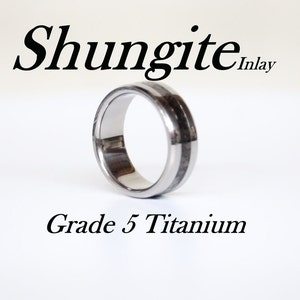 以下が含まれることがあります： 黒いシュンガイトのインレイが施された銀色のチタンリング。リングには「Shungite Inlay」と「Grade 5 Titanium」のラベルが貼られています。