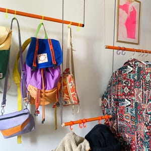 May include: A wall-mounted orange metal rack with hooks holds various bags, backpacks, and jackets. The rack is decorated with colorful straps and a bright orange and purple backpack with the text "Budo Chooses" printed on it. A patterned jacket hangs on a lower rack.