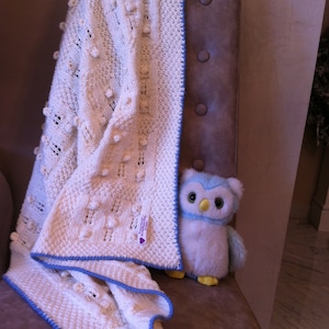May include: Cream-coloured knitted blanket with a blue border and textured details. A plush blue and white owl toy sits beside the blanket. The blanket has a tag with text.