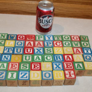 May include: A collection of wooden alphabet blocks in various colors including red, blue, green, and yellow, each featuring embossed letters. A 12 fluid ounce aluminum can of Busch Light Apple is positioned behind the grid of blocks for scale.
