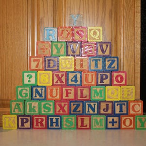 May include: A pyramid of wooden alphabet and number blocks in various colors including red, blue, green, and yellow, stacked next to a red Busch Light Apple aluminum beverage can. The blocks feature printed letters, numbers, and symbols.