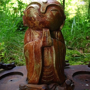 Peut inclure: Une petite statue en bois d'une personne avec un visage et des mains dorés, portant une robe brune. La statue est posée sur une base en bois avec un motif sculpté.