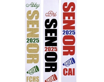 May include: Three white graduation sashes with gold, black, and red lettering. The sashes say "Senior 2025" with the names Abby, Bri, and Callie above the text. Each sash has a cheerleading megaphone with the school initials FCHS, NAHS, and CAI.