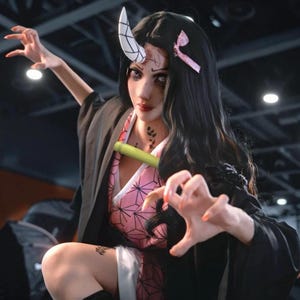 Op de afbeelding: Cosplay van een anime-personage. Het personage heeft lang zwart haar, een roze kimono met een geometrisch patroon en een zwarte jas. Ze heeft een bamboe buis in haar mond en een witte hoorn op haar hoofd.