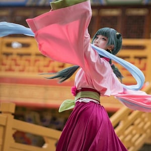 May include: A person in a traditional Asian costume with a pink top and a long, flowing magenta skirt. The costume includes a green and red belt, and blue and pink fabric streamers. The person has teal hair and is in a dance pose.