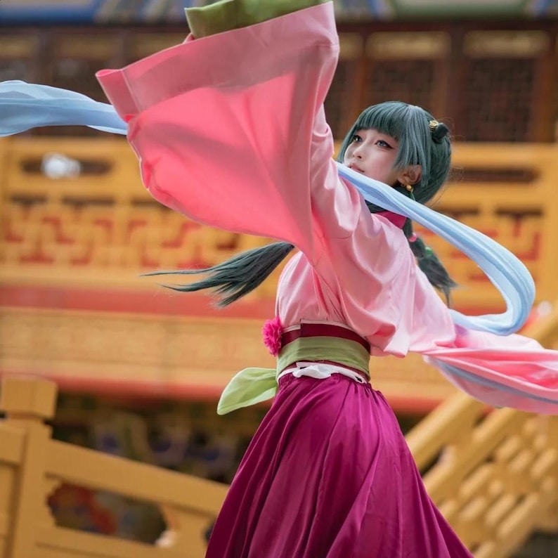 May include: A person in a traditional Asian costume with a pink top and a long, flowing magenta skirt. The costume includes a green and red belt, and blue and pink fabric streamers. The person has teal hair and is in a dance pose.