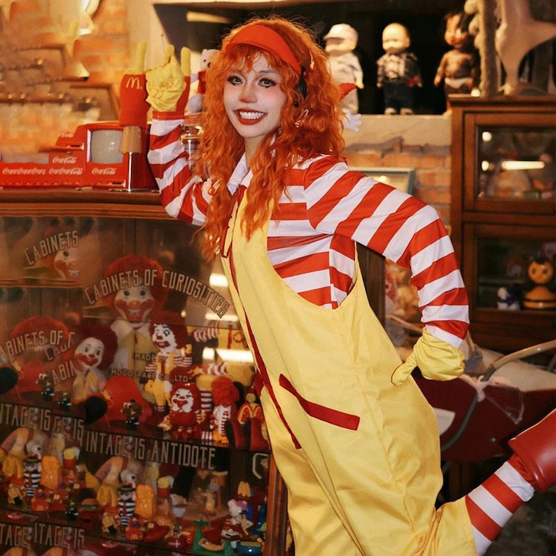 Puede incluir: Una persona disfrazada de Ronald McDonald, con un mono amarillo con detalles rojos, una camisa a rayas rojas y blancas y botas rojas. La persona sonr&iacute;e y lleva una peluca y una diadema rojas. El fondo incluye recuerdos vintage de Coca-Cola y una vitrina con mu&ntilde;ecos de payaso.
