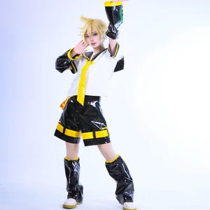 Può includere: Un costume cosplay con un top bianco con dettagli neri e una cravatta gialla, abbinato a pantaloncini e stivali neri con finiture gialle. L'outfit è completato da polsini neri e gialli. La persona indossa una parrucca bionda.