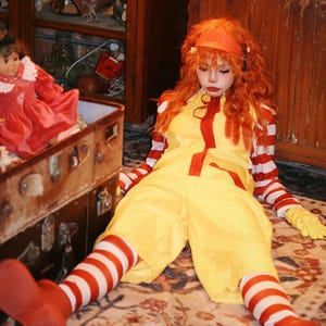 Puede incluir: Una persona con un mono amarillo con detalles rojos, mangas y calcetines a rayas rojas y blancas, y zapatos rojos. La persona tambi&eacute;n lleva una diadema naranja y tiene el pelo naranja. Una mu&ntilde;eca con un vestido rojo est&aacute; en una maleta vintage.
