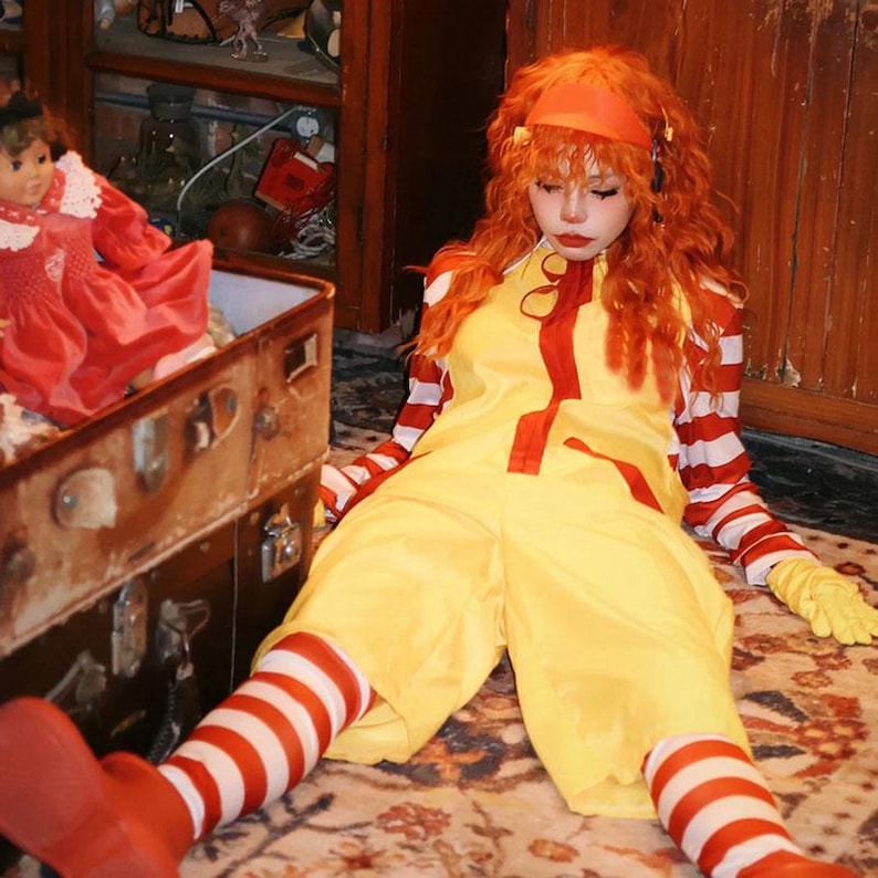 Puede incluir: Una persona con un mono amarillo con detalles rojos, mangas y calcetines a rayas rojas y blancas, y zapatos rojos. La persona tambi&eacute;n lleva una diadema naranja y tiene el pelo naranja. Una mu&ntilde;eca con un vestido rojo est&aacute; en una maleta vintage.