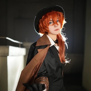 Puede incluir: Una persona con un traje negro con un abrigo marrón, un sombrero negro y una peluca naranja. La persona tiene ojos azules y lleva una camisa blanca con cuello y una gargantilla negra. El atuendo es un disfraz de cosplay.