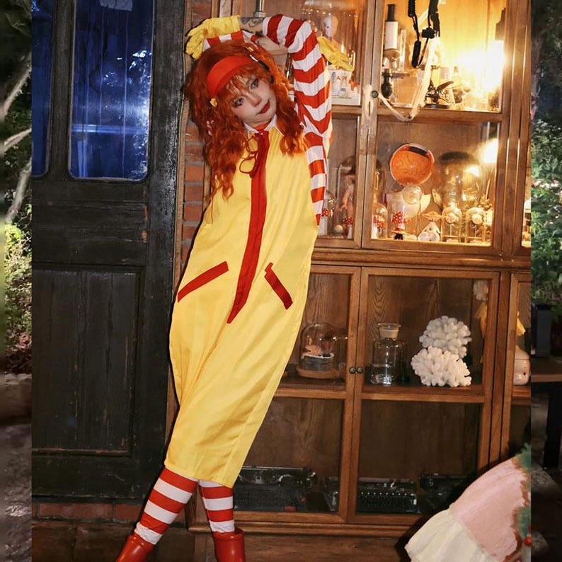 Puede incluir: Un mono amarillo con detalles rojos, que se asemeja a un disfraz de mascota de comida r&aacute;pida. El atuendo incluye mangas y calcetines a rayas rojas y blancas, combinados con botas rojas. Una diadema y una peluca rojas completan el look. El disfraz es una versi&oacute;n l&uacute;dica.