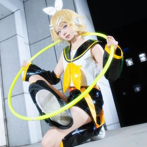 Può includere: Una persona in un costume cosplay, con un top bianco con colletto nero e dettagli gialli, abbinato a pantaloncini e maniche neri. L'outfit include un fiocco giallo e un cerchio giallo. La persona ha i capelli biondi e indossa accessori.