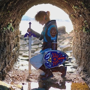 May include: A person dressed as Link from The Legend of Zelda, kneeling in a stone tunnel. They wear a blue and brown tunic, brown boots, and hold a Master Sword and a blue shield with a Triforce emblem.