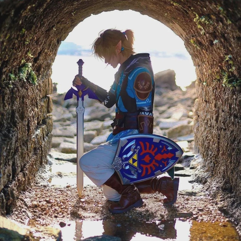 May include: A person dressed as Link from The Legend of Zelda, kneeling in a stone tunnel. They wear a blue and brown tunic, brown boots, and hold a Master Sword and a blue shield with a Triforce emblem.