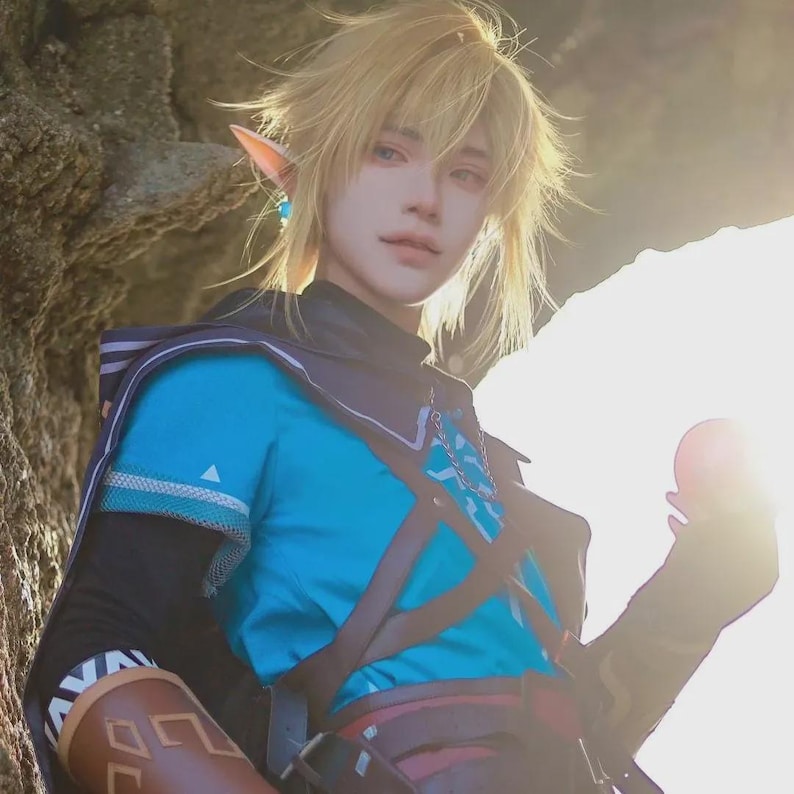 May include: A cosplay of a character with blonde hair, pointed ears, and blue eyes. The character wears a blue tunic with black accents, a brown leather harness, and a dark cape. The costume is set against a natural background with sunlight.