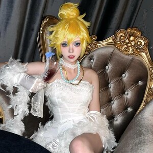 Può includere: Una persona con un abito bianco senza spalline con dettagli in pizzo, un boa di piume e guanti, che tiene in mano un bicchiere di liquido rosso. Indossa una parrucca gialla, occhi blu e una collana turchese, seduta su una sedia dorata ornata.