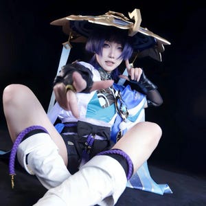 May include: A person wearing a cosplay outfit with a large hat, white and blue top, and black shorts. The outfit includes white boots with purple accents. The person is pointing towards the viewer.