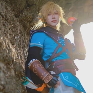 May include: A cosplay outfit inspired by a video game character. The costume features a blue tunic with brown accents, a brown leather-like belt, and a matching arm guard. The character has blonde hair and pointed ears, holding a small, round object.