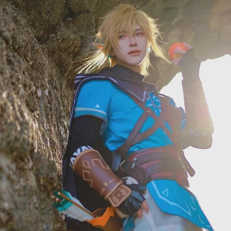 May include: A cosplay outfit inspired by a video game character. The costume features a blue tunic with brown accents, a brown leather-like belt, and a matching arm guard. The character has blonde hair and pointed ears, holding a small, round object.