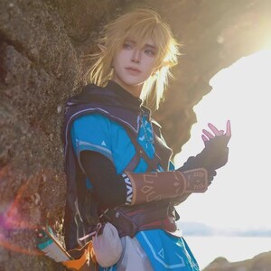 May include: A cosplay outfit inspired by a video game character. The costume features a blue tunic with brown accents, a black hood, and a belt with pouches. The character has blonde hair and pointed ears, with a bow and arrow visible.
