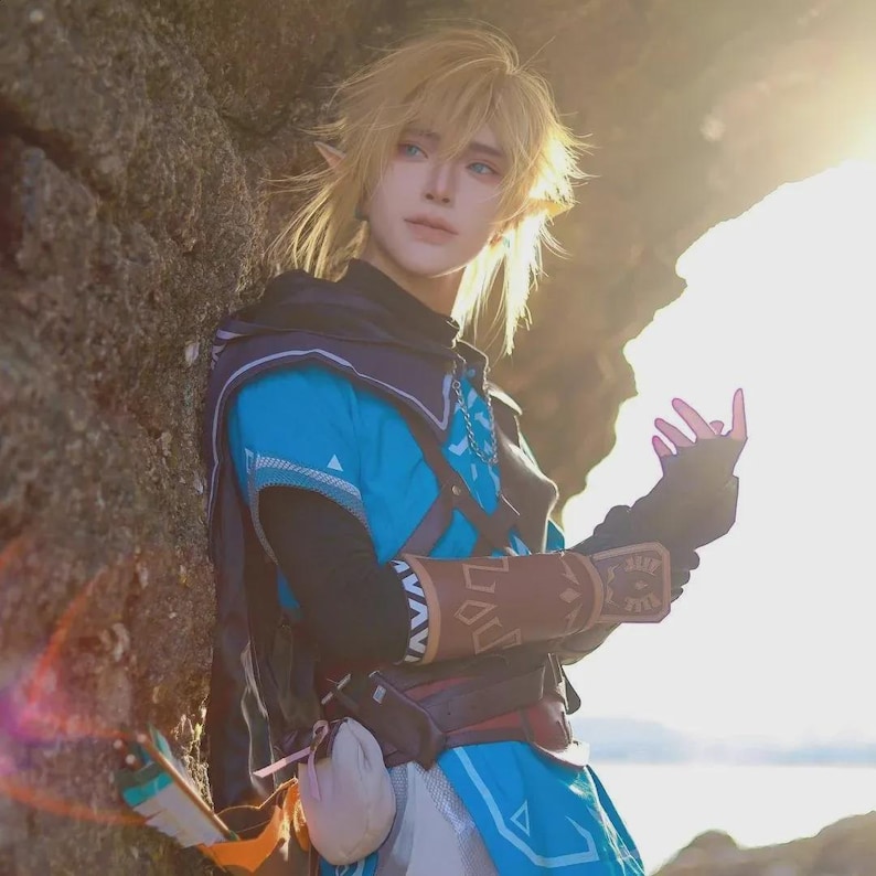 May include: A cosplay outfit inspired by a video game character. The costume features a blue tunic with brown accents, a black hood, and a belt with pouches. The character has blonde hair and pointed ears, with a bow and arrow visible.