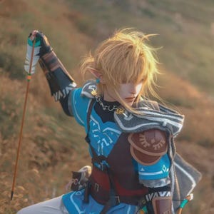 May include: A cosplay of a character from The Legend of Zelda. The costume features a blue tunic with white accents, brown leather armor, and a gray cape. The character is holding a bow and arrow with a feathered end. The background is a grassy field.