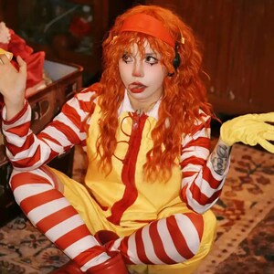 Puede incluir: Una persona con un disfraz amarillo y rojo con mangas y leggings a rayas rojas y blancas. El disfraz tiene una cremallera roja y una diadema roja. La persona lleva guantes amarillos y tiene el pelo y maquillaje naranja.