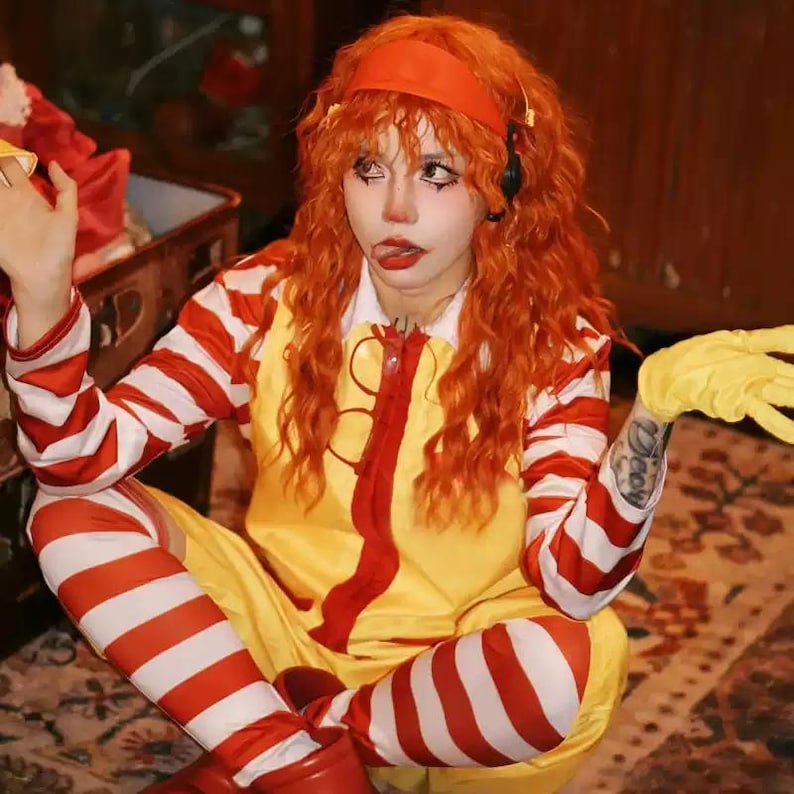 Puede incluir: Una persona con un disfraz amarillo y rojo con mangas y leggings a rayas rojas y blancas. El disfraz tiene una cremallera roja y una diadema roja. La persona lleva guantes amarillos y tiene el pelo y maquillaje naranja.