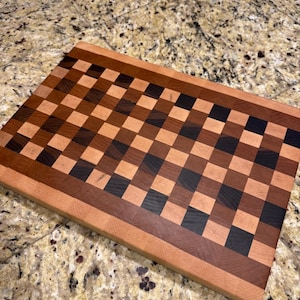 May include: A rectangular wooden cutting board with a checkerboard pattern. The board features alternating squares of light, medium, and dark brown wood, with a border of lighter wood. The board is approximately 18 inches long and 12 inches wide.
