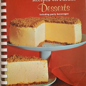 May include: A spiral-bound cookbook titled Recipes on Parade Desserts including party beverages. The cover features a yellow cheesecake with a brown crumb topping on a light blue cake stand, with text stating 2000 WORLD WIDE FAVORITES OF MILITARY OFFICERS' WIVES.