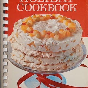 May include: A spiral-bound cookbook titled The Officers' Wives Holiday Cookbook with a red, white, and blue cover. The front features a photograph of a multi-layered cake topped with diced fruit, sitting on a glass stand with a red ribbon underneath.
