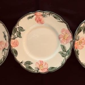 May include: Three white ceramic plates with a floral design. The plates have a scalloped edge and feature pink roses with green leaves.