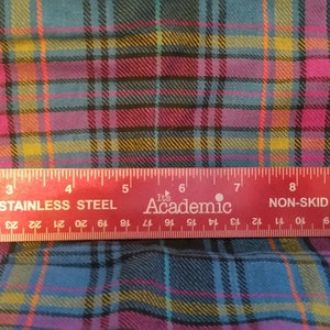 Könnte beinhalten: Nahaufnahme eines karierten Stoffes mit einem Lineal, das 28 cm misst. Der Stoff ist eine Mischung aus Rosa, Blau, Gelb und Schwarz.