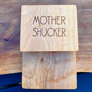May include: A light-colored wooden coaster with the words "MOTHER SHUCKER" engraved on the top. The coaster is square-shaped and sits on a wooden surface. The wood grain is visible.