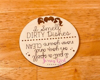 May include: A round wooden sign with the text "I Smell DIRTY Dishes" and "I put a spell on you and now you're CLEAN". The sign has a graphic of three witch silhouettes and the words "Jersey Girl GRAPHICS".
