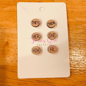 May include: Six wooden round tags with the words "They", "Them", "Her", "She", "Him", and "He" printed in black.