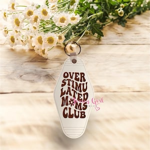 以下が含まれることがあります： 茶色の文字で「OVER STIMULATED MOMS CLUB」と書かれた白いモーテル風キーホルダー。キーホルダーはシルバーのリングに付いています。背景には白い花と緑があります。