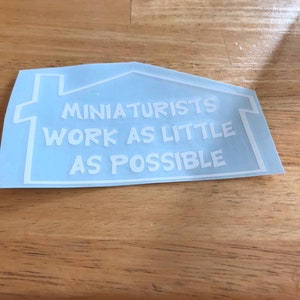 May include: A light blue sticker with a white outline of a house. The text inside the house reads "Miniaturists work as little as possible."