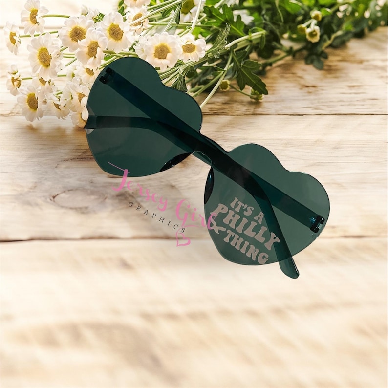 May include: Heart-shaped sunglasses with dark green lenses and frames. The right lens features the text "It's a Philly Thing" in white. The sunglasses are resting on a light wooden surface, with white flowers in the background.