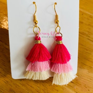 May include: A pair of pink, white, and cream tassel earrings with gold hooks. The tassels are layered and have a fringed edge.