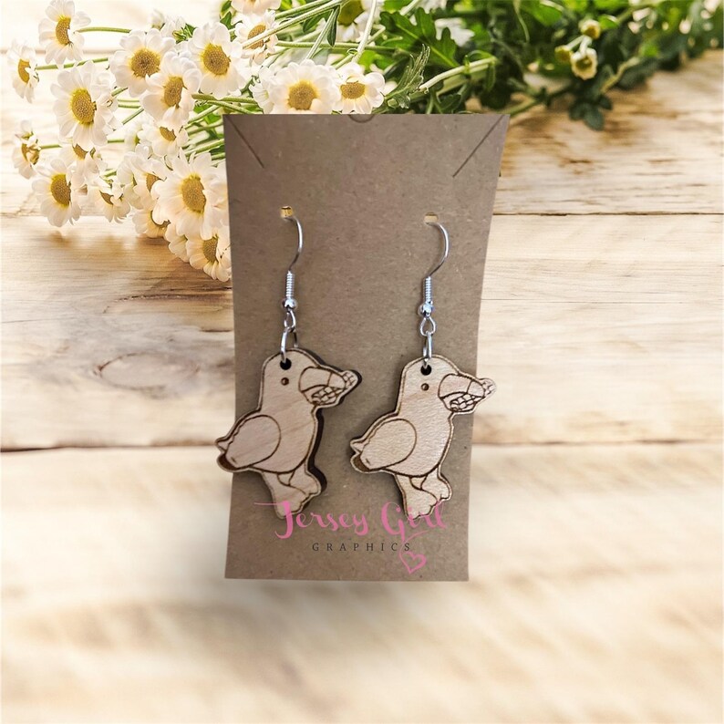 May include: A pair of wooden earrings featuring a cartoon bird design. The earrings are on a brown card with the text "Jersey Girl Graphics".