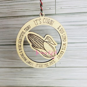 Può includere: Un ornamento di legno a forma di cerchio con una pannocchia di mais all'interno. Il testo sull'ornamento recita "Non riesco a immaginare qualcosa di più bello. È mais! Un grosso grumo con nodi. Ha il succo."