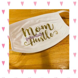 May include: White face mask with gold glitter lettering that says "Mom Hustle" and a small purple text that says "GeekEssentials".