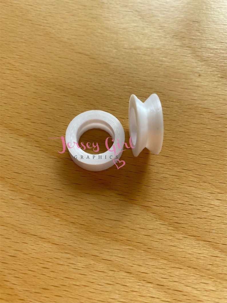 May include: Two white, spool-shaped objects are displayed on a light brown wooden surface. One is a complete spool, and the other is standing upright. The text "Jersey Girl Graphics" is visible on the spool.