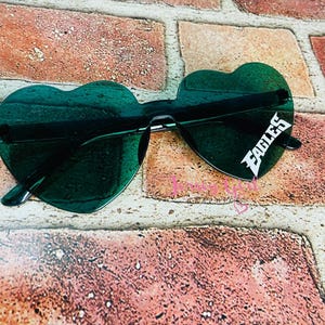 May include: Heart-shaped sunglasses with dark green lenses. The sunglasses have a black frame and the word "EAGLES" in white on the right lens. The sunglasses are resting on a brick surface.