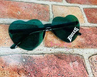 May include: Heart-shaped sunglasses with dark green lenses. The sunglasses have a black frame and the word "EAGLES" in white on the right lens. The sunglasses are resting on a brick surface.