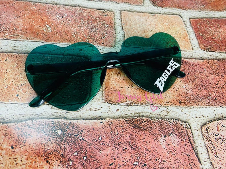 May include: Heart-shaped sunglasses with dark green lenses. The sunglasses have a black frame and the word "EAGLES" in white on the right lens. The sunglasses are resting on a brick surface.