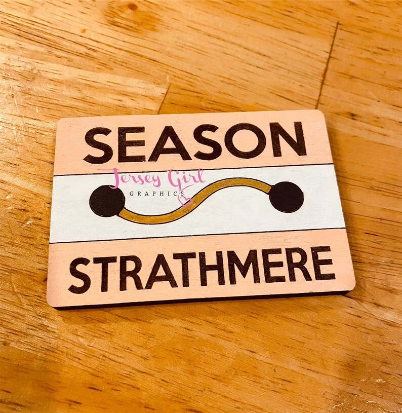 May include: A pink and white wooden sign with the text "SEASON" in black letters, a gold and black graphic of a curved line with two black circles at the ends, and the text "STRATHMERE" in black letters.